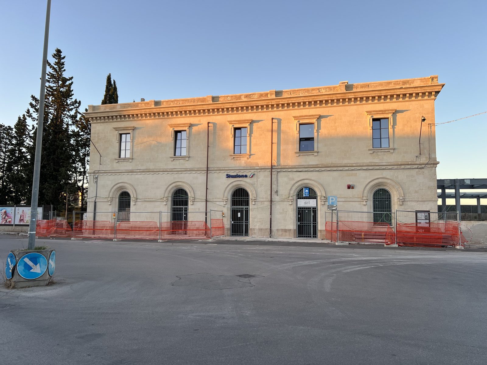 La stazione ferroviaria di Manduria al tramonto, il fabbricato viaggiatori in pietra del 1907 con l’insegna Stazione, le reti arancioni del cantiere davanti e la nuova pensilina sulla destra