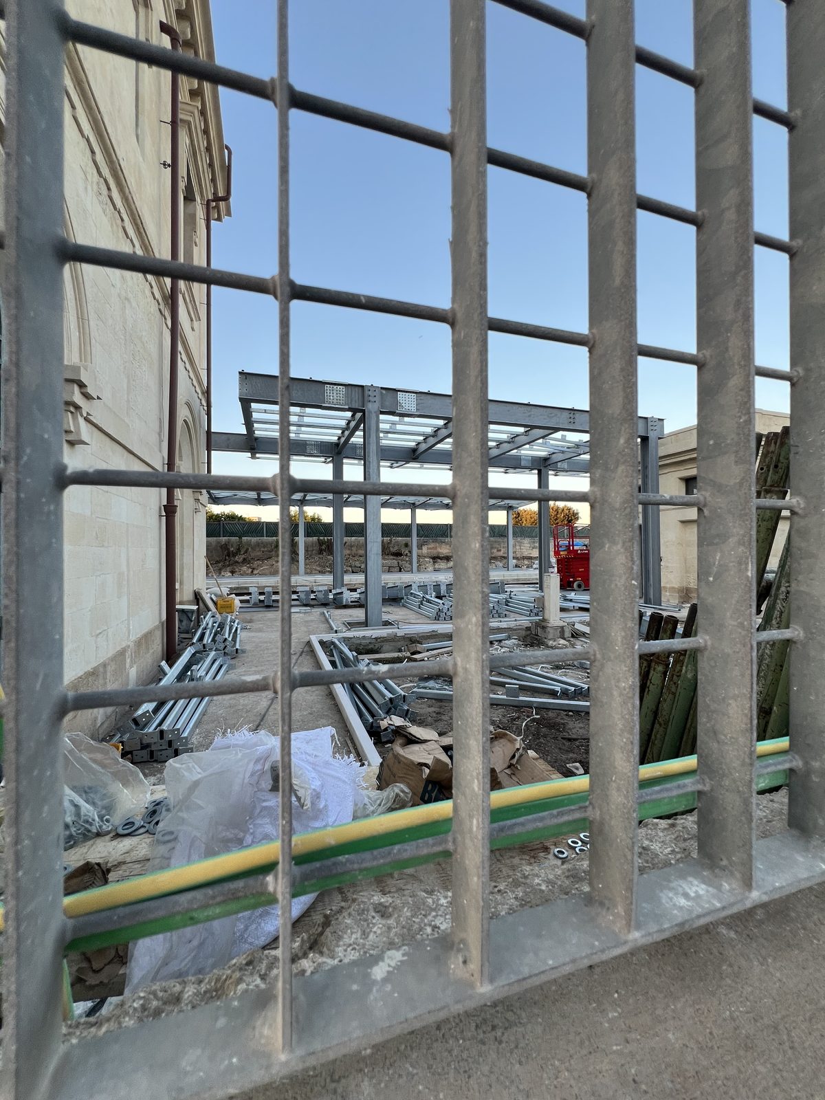 Lo scheletro in acciaio della nuova pensilina della stazione di Manduria in costruzione, visto attraverso una grata, con i profilati accatastati a terra
